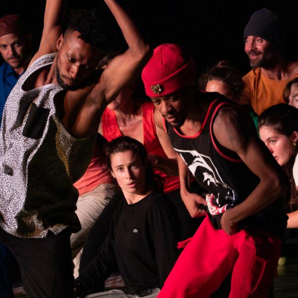 Des danseurs et danseuses dans une pièce sombre sont en plein mouvement. Un danseur se détache du groupe pour un solo, les deux bras et une jambe levés, tandis que le reste se tient en arrière, accroupis.
