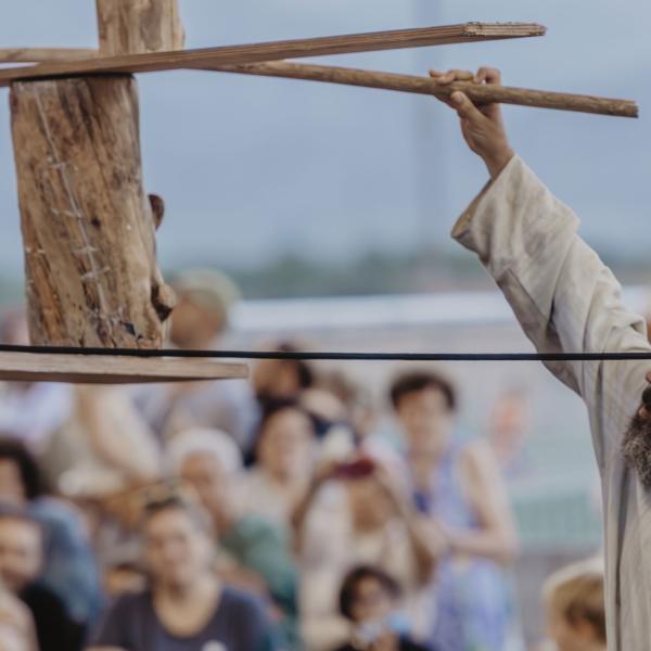 L'artiste au premier plan tient dans une main une corde reliée à une structure en bois en équilibre, dont il tient une extrémité de l'autre main. Le public à l'arrière-plan est flou.