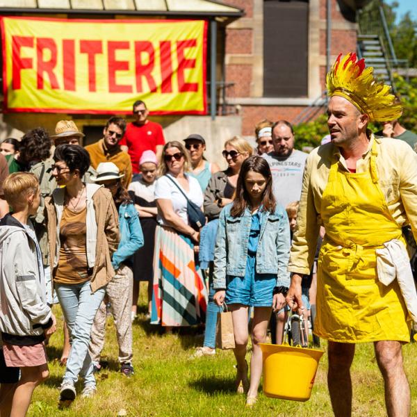 Un homme dans un jardin vêtu d'une couronne de plumes rouges et jaunes ainsi que d'une chemise et d'un tablier jaunes porte dans chaque main un seau jaune rempli de pommes de terres. Il est suivi par une trentaine de personnes de tout âges. Au fond, une grande bannière "FRITERIE" écrit rouge sur jaune est accrochée sur un bâtiment.