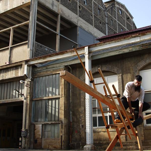 L'artiste se tient en équilibre sur une construction en bois. En arrière-plan, un bâtiment industriel abandonné.