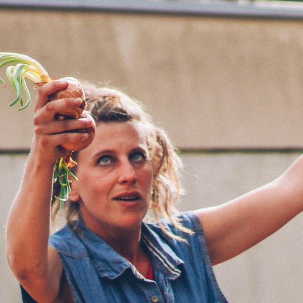 La comédienne a les deux bras levés et tient dans chaque main deux oignons. Elle porte son regard vers sa main droite. 