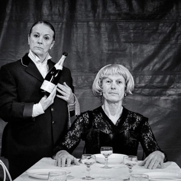 Photo en noir et blanc. Le comédien, habillé en bourgeoise, est assis à une table préparée pour un dîner. La comédienne est debout à gauche, habillée en majordome, et tient une bouteille de champagne.
