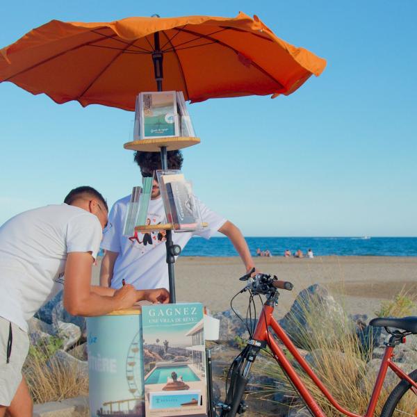 Au bord de la plage, l'artiste se tient derrière son triporteur publicitaire. L'avant du vélo est constitué d'un parasol orange qui porte des flyers et d'un support en bois. Un homme est en train d'écrire, appuyé sur le plateau en bois.