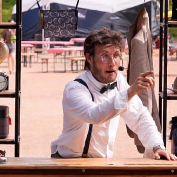 Le comédien est debout derrière un haut bureau en bois, dehors. Habillé d'une chemise blanche, nœud papillon noir et bretelles, il porte un micro serre-tête et pointe le public du doigt. Derrière lui, des étagères sont remplies de bibelots, boîtes et objets de différentes tailles.
