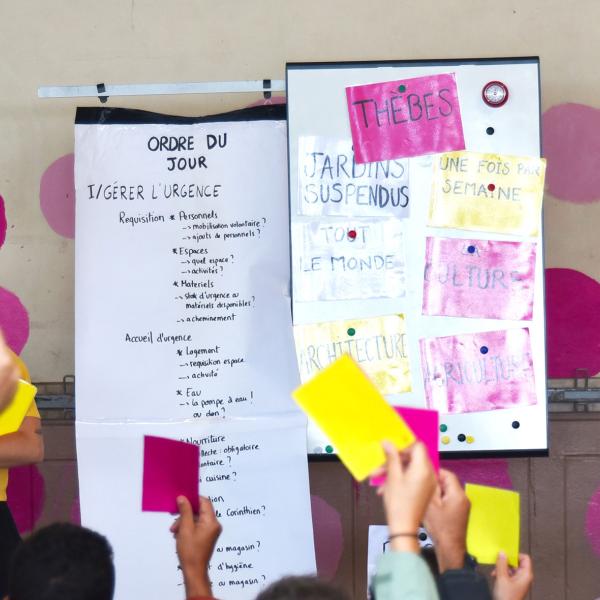 Au centre, deux panneaux blancs : l'un avec "L'ordre du jour", l'autre avec des pancartes colorées "Thèbes", "Jardins suspendus"... À gauche, la comédienne porte un t-shirt jaune et une pancarte jaune "Annulées". À droite, le comédien porte un t-shirt rose et une pancarte rose "Déplacées à l'extérieur". Le public brandit des cartons jaunes ou roses.