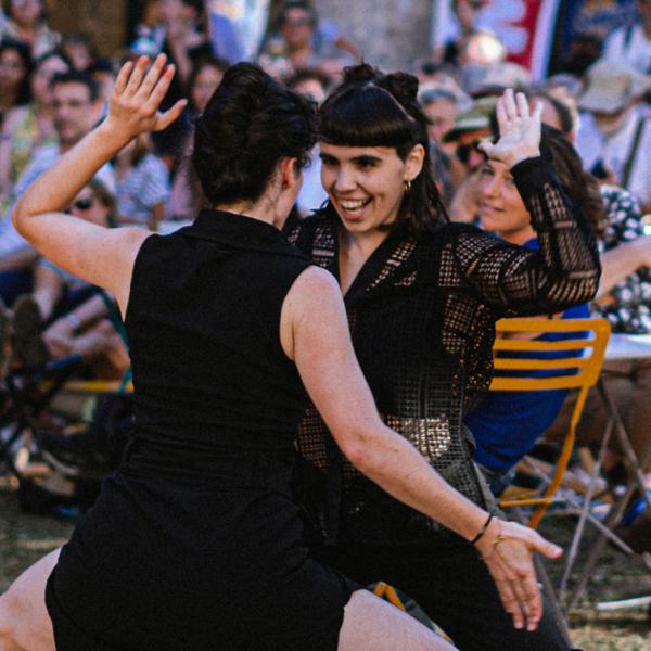 Deux danseuses sont au milieu de personnes assises à des tables en extérieur. Elles se font face, accroupies et l'une sourit. Elles ont une main en l'air et l'autre en bas.
