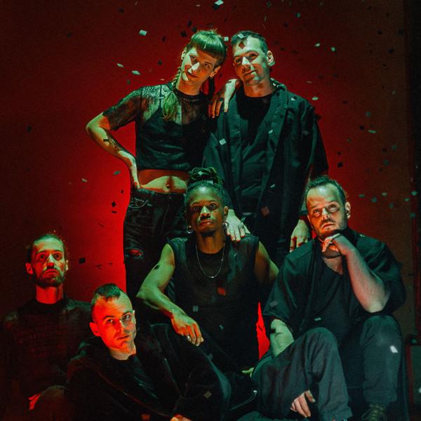 Les six membres du groupe posent devant un fond sombre éclairé en rouge. Des paillettes tombent. Deux sont debout, deux à genoux, un assis et un allongé.