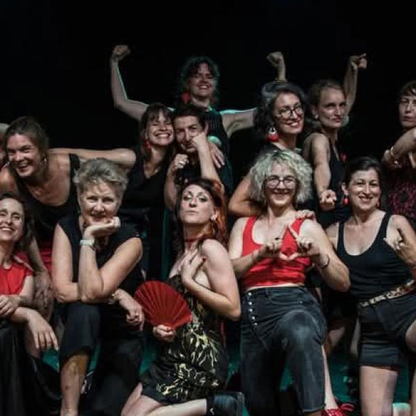 Les membres de la chorale posent sur une scène en intérieur. La pièce est sombre. Elles portent du rouge et/ou du noir. Certaines font des signes avec les mains ou lèvent les bras.