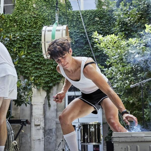 Sur une scène dans une cour intérieure, une artiste allume un fumigène tandis qu'un musicien et une musicienne jouent de la batterie sur le côté. Un tom de batterie est accroché en l'air.