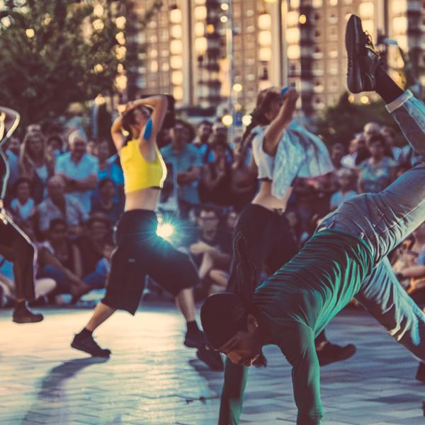 Une troupe de danseuses et danseurs en mouvement, encerclés par le public en extérieur. Un danseur à l'avant a deux mains et un pied à terre, et une jambe pliée en l'air. À l'arrière, cinq membres de la troupe avancent en diagonale en se couvrant la tête.