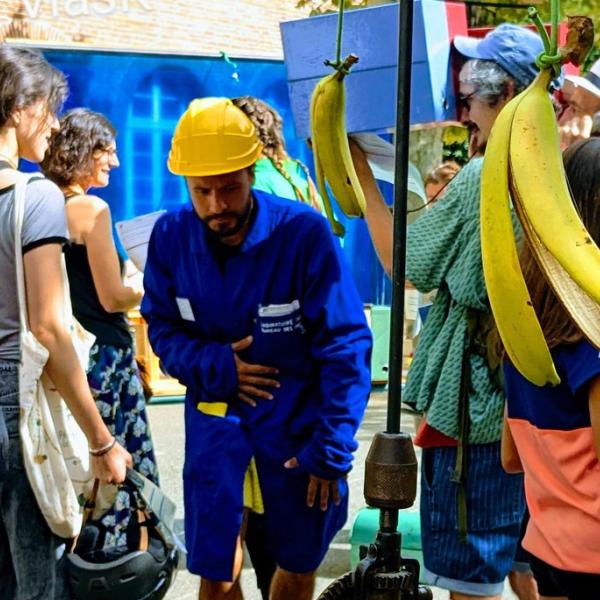 Des personnes dehors, un garçon tient une cannes à pêche avec des bananes au bout. Au milieu se tient un comédien avec une blouse bleue et un casque de chantier.