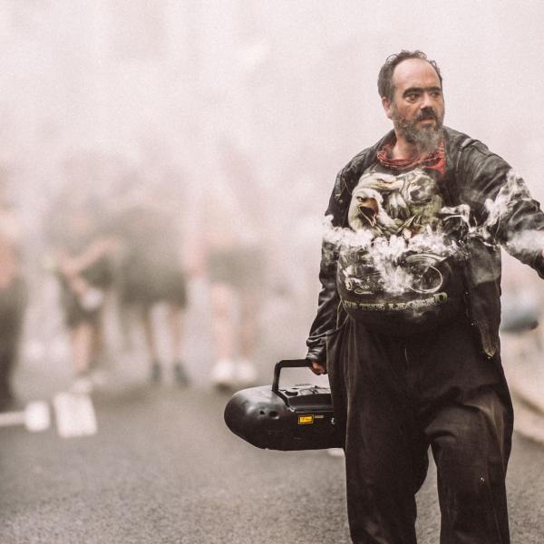 Un homme se tient au milieu de la rue. Il tient dans une main une grosse radio CD et dans l'autre un fumigène blanc. La rue est enfumée et un groupe de personne du public se tient à l'arrière, dans la fumée.