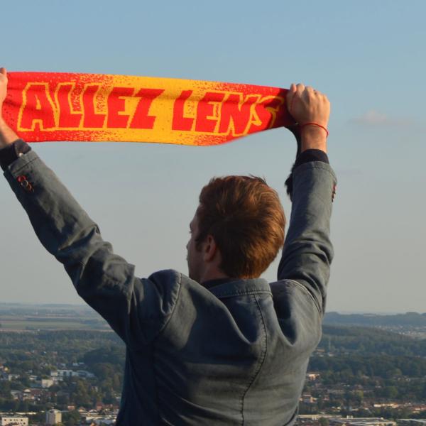 Le comédien se tient de dos et brandit une écharpe de foot rouge et jaune "ALLEZ LENS". Il regarde l'horizon et les villes environnantes en contrebas.