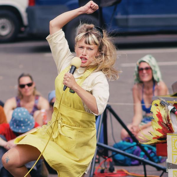 La comédienne porte un tablier jaune et tient un micro dans sa main gauche. Elle semble en train de sauter, un bras en l'air et une jambe pliée. Derrière elle, un coupe-frites ambulant, décoré avec des motifs jaunes et l'inscription "Friterie". À l'arrière, le public est assis par terre, légèrement flouté.