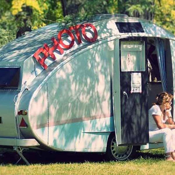 Caravane vintage dans une étendue d'herbe avec un lettrage "PHOTO" posé dessus. Une femme est assise sur le rebord de la porte.