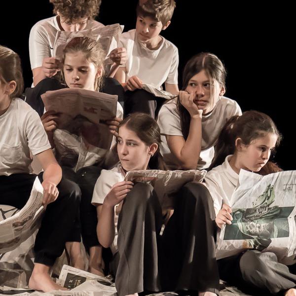 Neufs enfants sont assis et assises sur scène et lisent des journaux. Iels portent un t-shirt blanc et un pantalon noir. Des journaux sont éparpillés sur le sol.