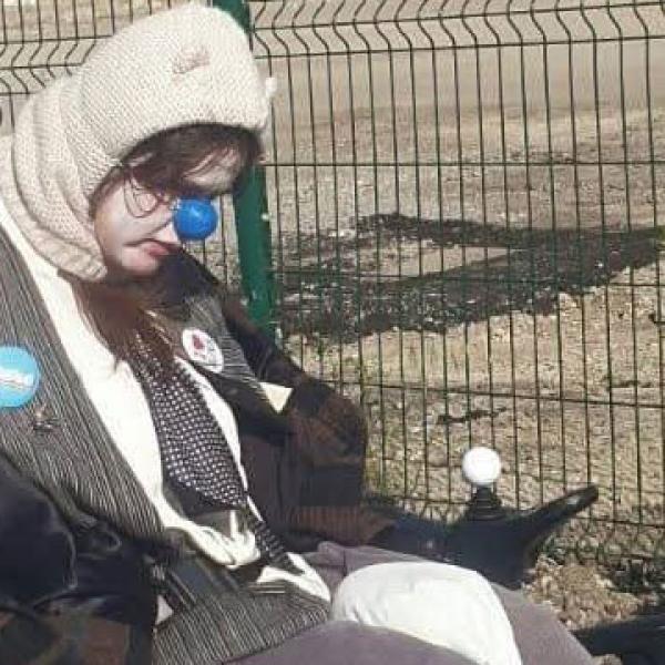 Un clown en fauteuil roulant électrique porte une cagoule blanche avec des petites oreilles. Son visage est visible, peint en blanc et il porte un nez bleu en plastique. Au dos de son fauteuil sont accrochées des pancartes en carton.