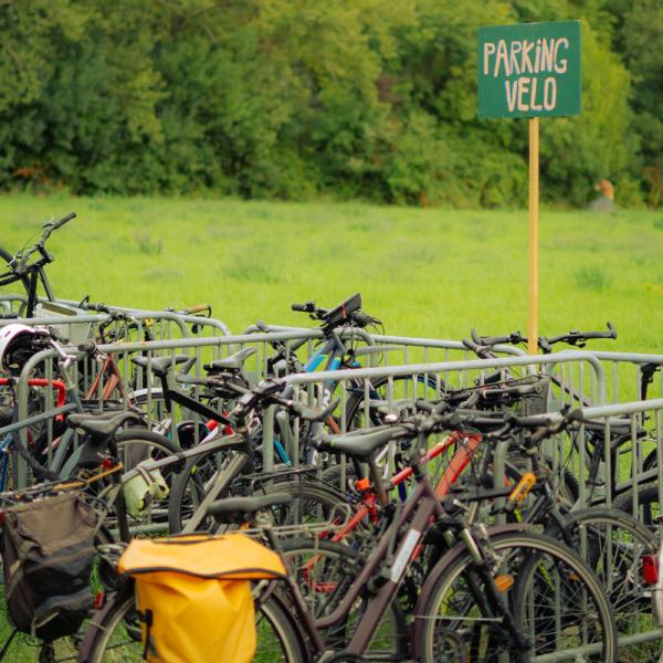 Dans un champ, des vélos sont parqués, attachés à des barrières vauban. Une pancarte en bois colorée indique "PARKING VELO".
