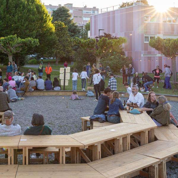 Installation en bois de tables et bancs installés en octogone dans la Cité Rose/Résidence St. Agne de Ramonville. Des personnes sont assises et des enfants jouent autour pendant un concert.