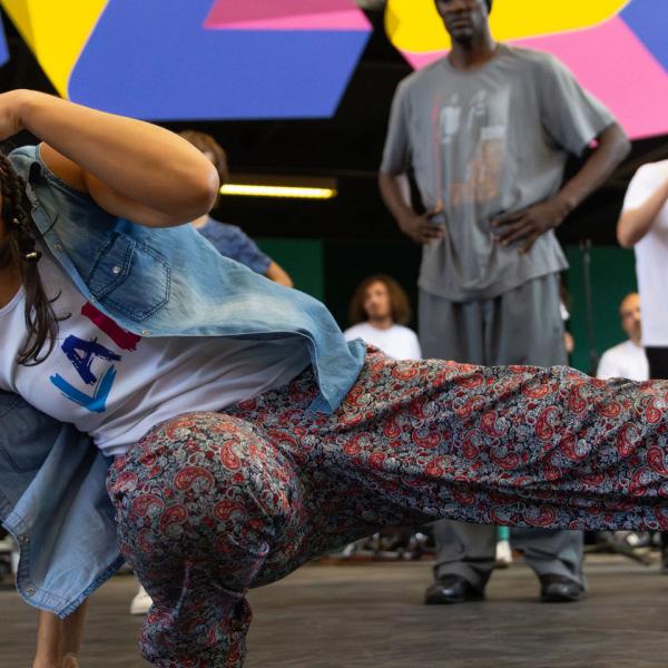 Une danseuse en plein mouvement de hip-hop. Elle a une main au sol, une jambe pliée et l'autre tendue vers le côté. Sa deuxième main couvre son front. Des personnes la soutiennent autour.