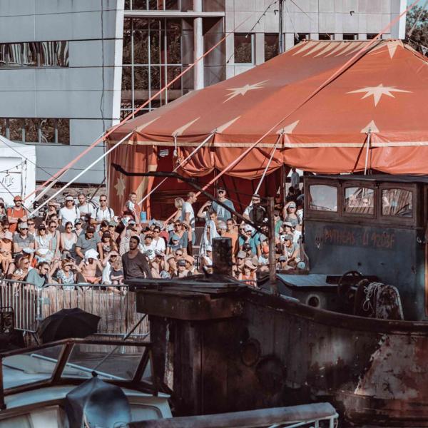 Photo d'une édition précédente du festival. Côté place du canal, les bateaux sont amarrés et le public se tient debout à côté d'un grand barnum rouge. L'entrée de l'espace est défini par une énorme arche en carton décorée de dessins avec écrit ARTO.