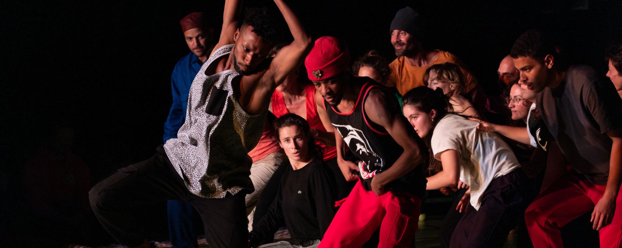 Des danseurs et danseuses dans une pièce sombre sont en plein mouvement. Un danseur se détache du groupe pour un solo, les deux bras et une jambe levés, tandis que le reste se tient en arrière, accroupis.