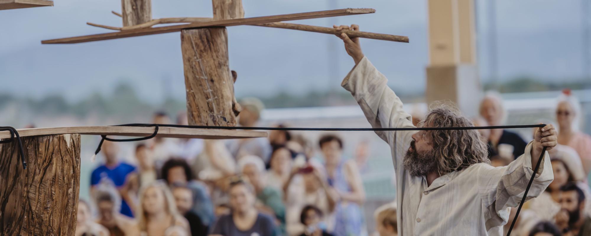 © Cia Nacho Flores L'artiste au premier plan tient dans une main une corde reliée à une structure en bois en équilibre, dont il tient une extrémité de l'autre main. Le public à l'arrière-plan est flou.