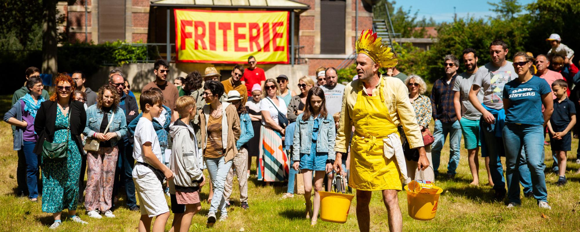 © Kalimba Un homme dans un jardin vêtu d'une couronne de plumes rouges et jaunes ainsi que d'une chemise et d'un tablier jaunes porte dans chaque main un seau jaune rempli de pommes de terres. Il est suivi par une trentaine de personnes de tout âges. Au fond, une grande bannière "FRITERIE" écrit rouge sur jaune est accrochée sur un bâtiment.