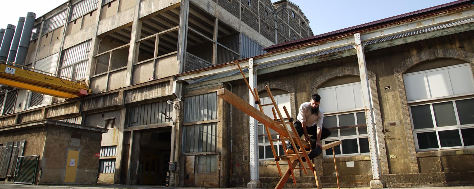 © Stéphane Rouaud L'artiste se tient en équilibre sur une construction en bois. En arrière-plan, un bâtiment industriel abandonné.
