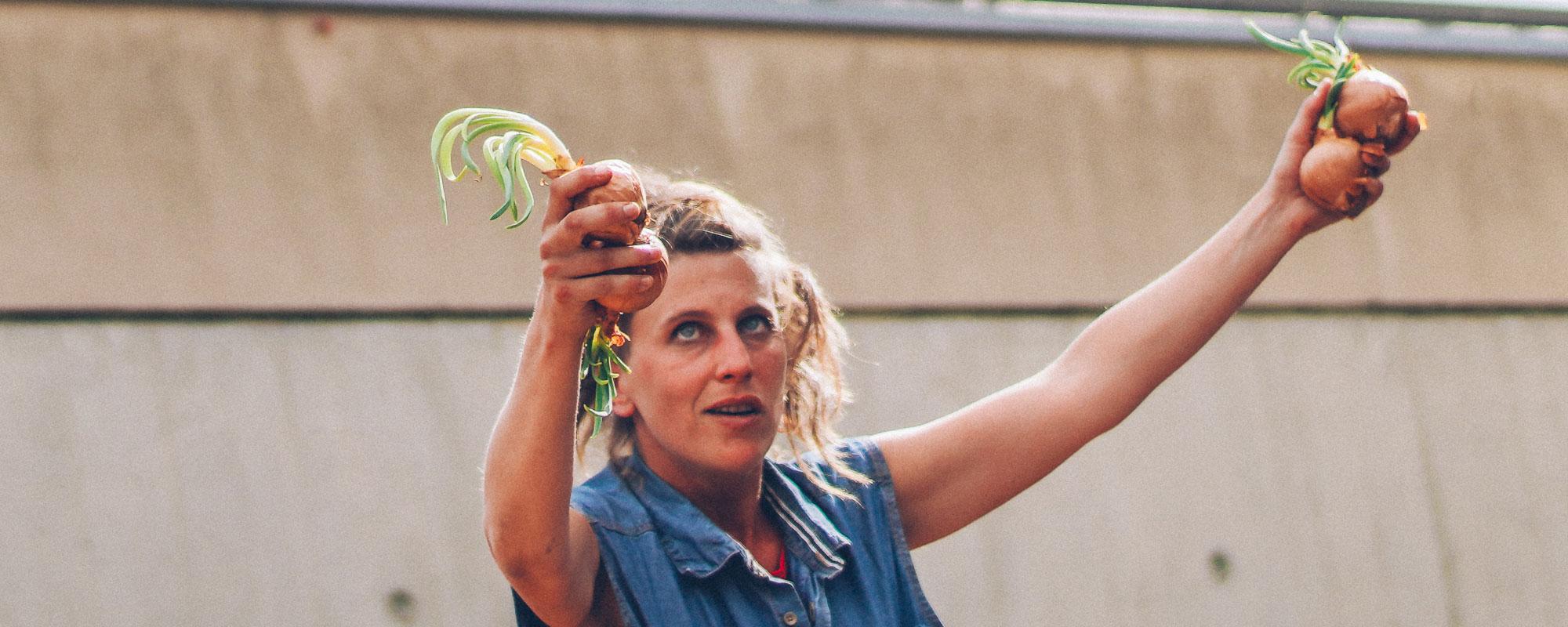 La comédienne a les deux bras levés et tient dans chaque main deux oignons. Elle porte son regard vers sa main droite.