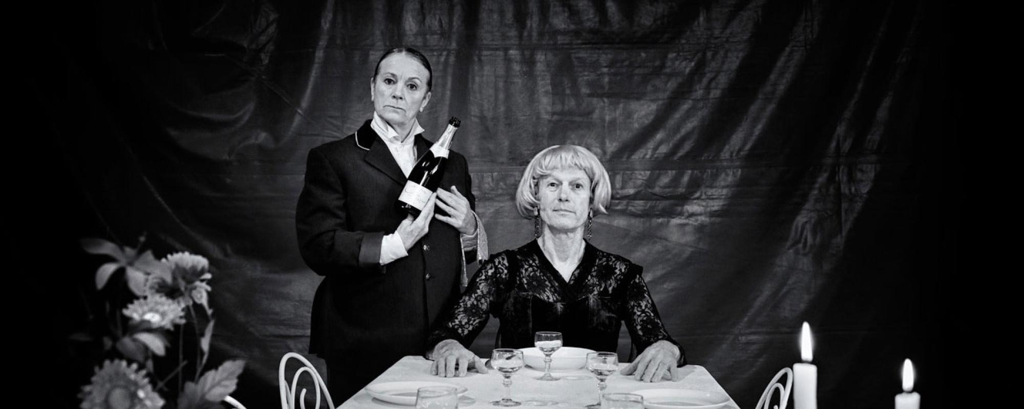 © Wilfried Thomas Photo en noir et blanc. Le comédien, habillé en bourgeoise, est assis à une table préparée pour un dîner. La comédienne est debout à gauche, habillée en majordome, et tient une bouteille de champagne.