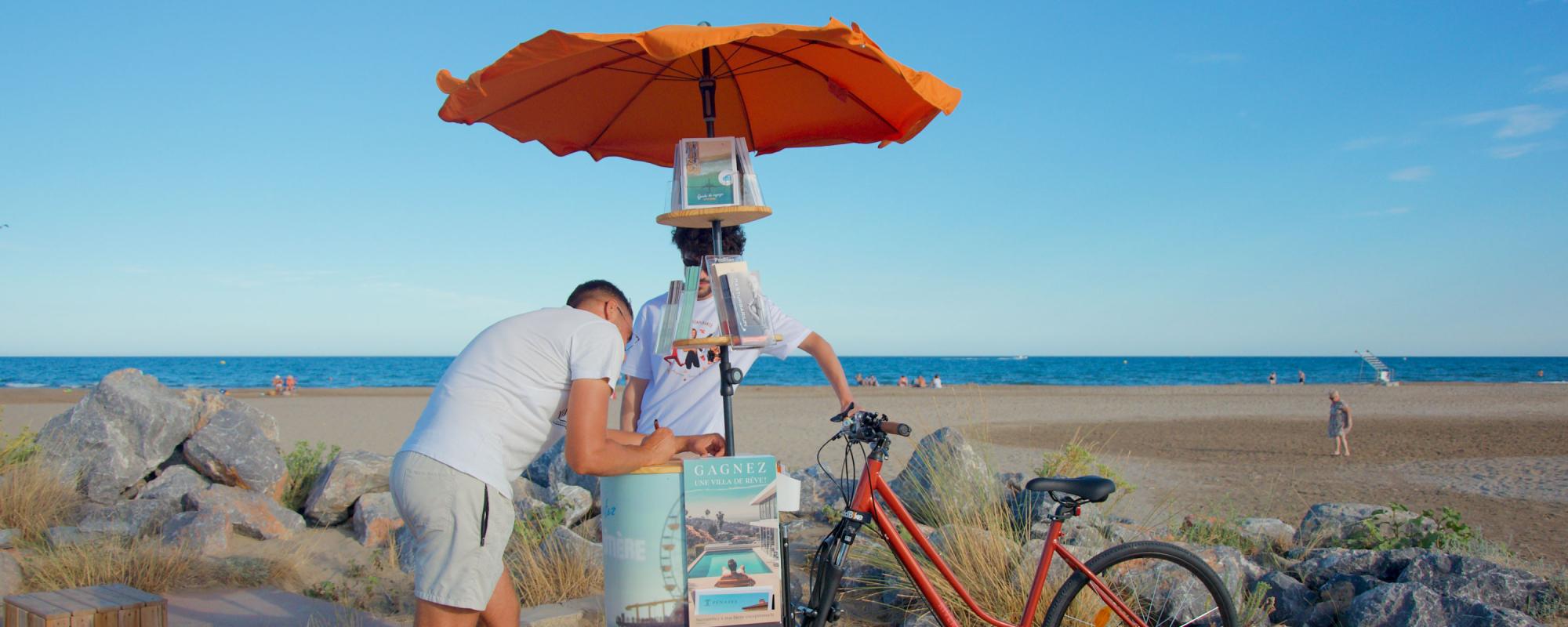 © Léa Lebrun Au bord de la plage, l'artiste se tient derrière son triporteur publicitaire. L'avant du vélo est constitué d'un parasol orange qui porte des flyers et d'un support en bois. Un homme est en train d'écrire, appuyé sur le plateau en bois.