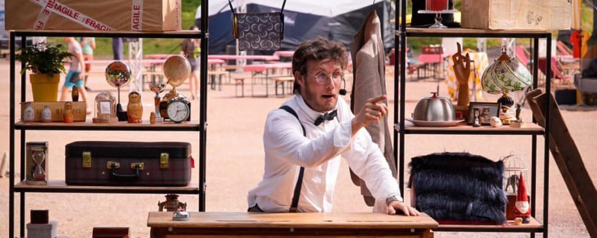 Le comédien est debout derrière un haut bureau en bois, dehors. Habillé d'une chemise blanche, nœud papillon noir et bretelles, il porte un micro serre-tête et pointe le public du doigt. Derrière lui, des étagères sont remplies de bibelots, boîtes et objets de différentes tailles.