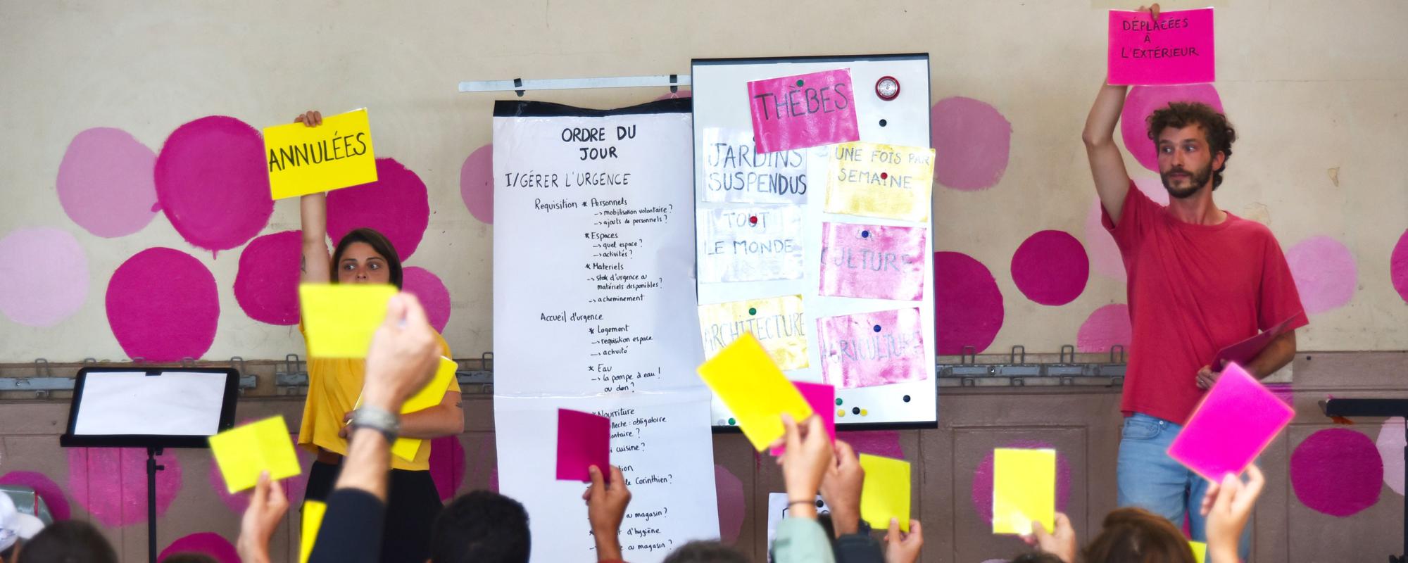 © Maxime Grimardias Au centre, deux panneaux blancs : l'un avec "L'ordre du jour", l'autre avec des pancartes colorées "Thèbes", "Jardins suspendus"... À gauche, la comédienne porte un t-shirt jaune et une pancarte jaune "Annulées". À droite, le comédien porte un t-shirt rose et une pancarte rose "Déplacées à l'extérieur". Le public brandit des cartons jaunes ou roses.