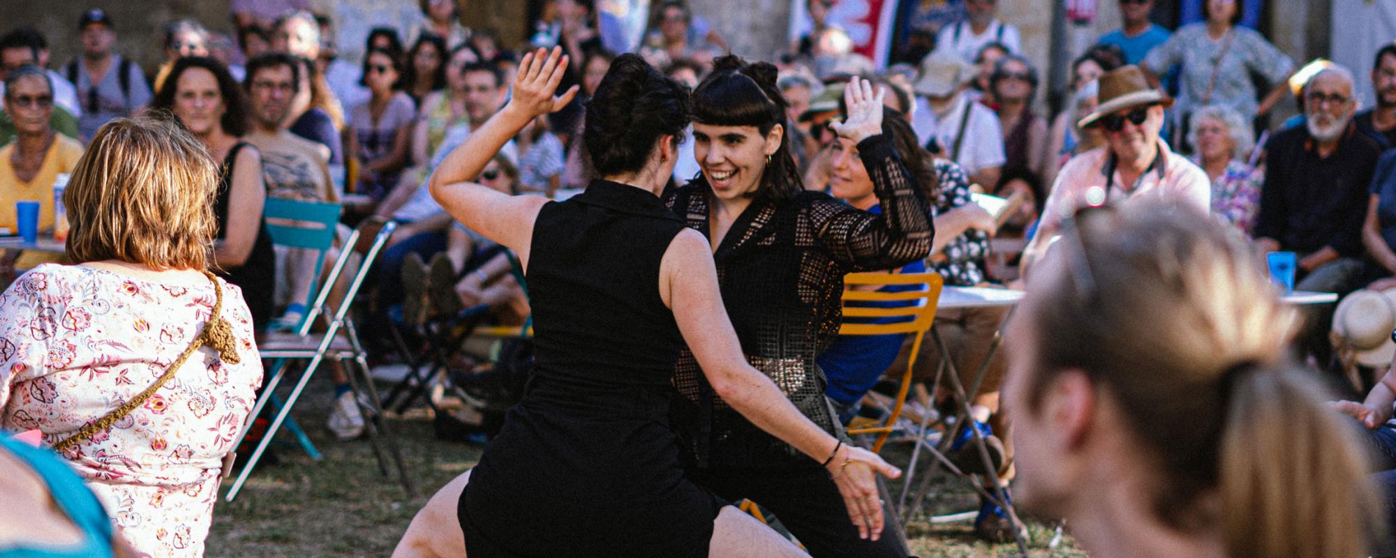 © Lucie Fournaise - Fournaise Factory Deux danseuses sont au milieu de personnes assises à des tables en extérieur. Elles se font face, accroupies et l'une sourit. Elles ont une main en l'air et l'autre en bas.