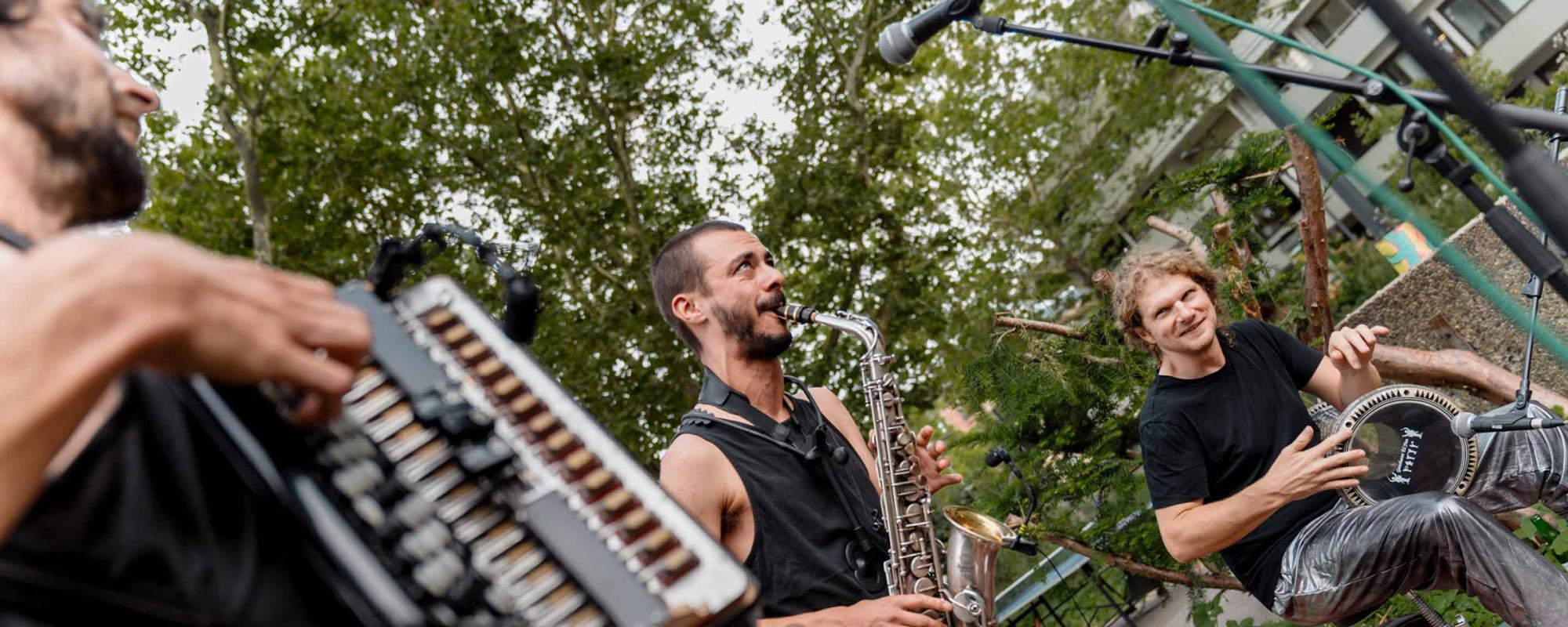 Trois musiciens en extérieur ; un joue de l'accordéon, un joue du saxophone et le dernier du darbuka.