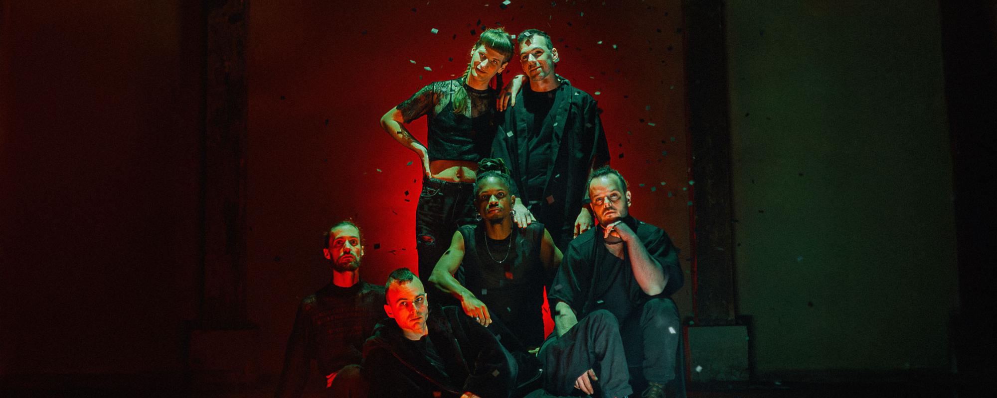 Les six membres du groupe posent devant un fond sombre éclairé en rouge. Des paillettes tombent. Deux sont debout, deux à genoux, un assis et un allongé.