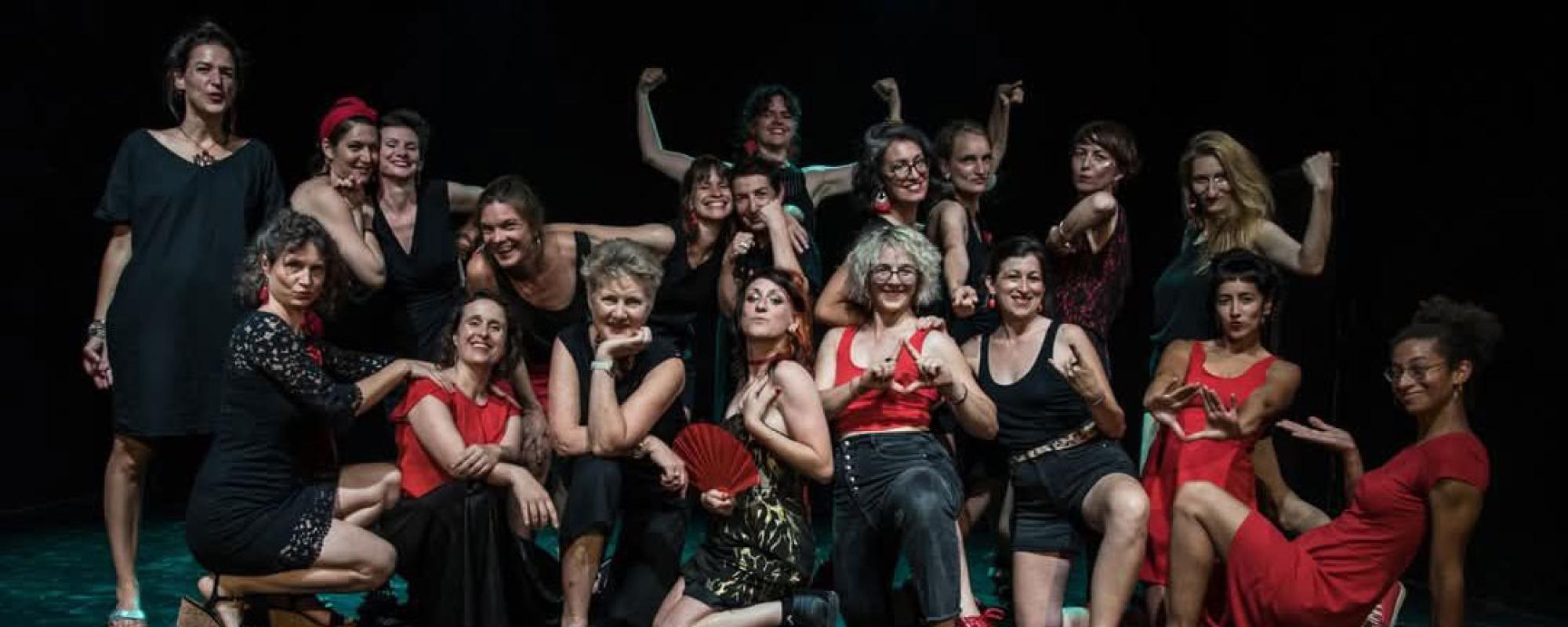 © Chorale de l'Ébranleuse Les membres de la chorale posent sur une scène en intérieur. La pièce est sombre. Elles portent du rouge et/ou du noir. Certaines font des signes avec les mains ou lèvent les bras.