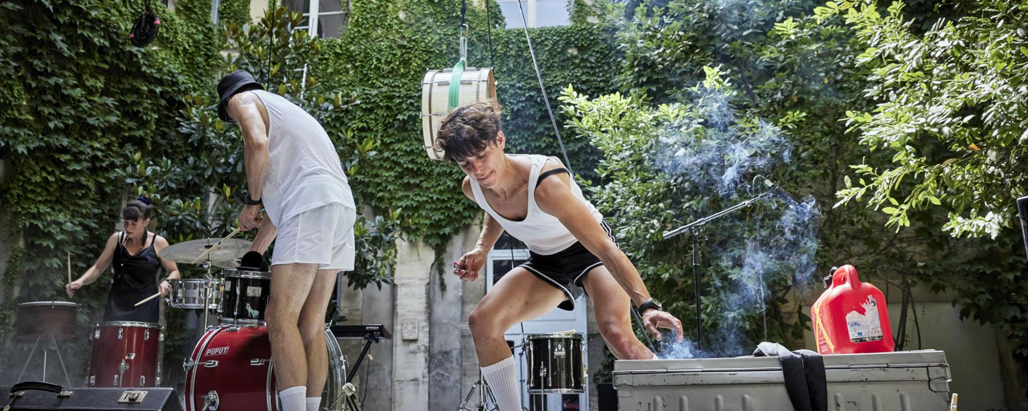 Sur une scène dans une cour intérieure, une artiste allume un fumigène tandis qu'un musicien et une musicienne jouent de la batterie sur le côté. Un tom de batterie est accroché en l'air.