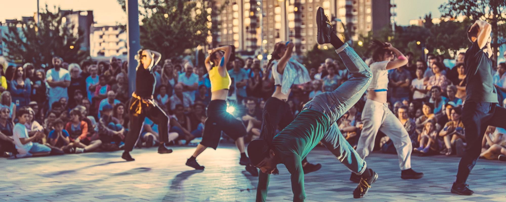 © Loïc Nys Une troupe de danseuses et danseurs en mouvement, encerclés par le public en extérieur. Un danseur à l'avant a deux mains et un pied à terre, et une jambe pliée en l'air. À l'arrière, cinq membres de la troupe avancent en diagonale en se couvrant la tête.