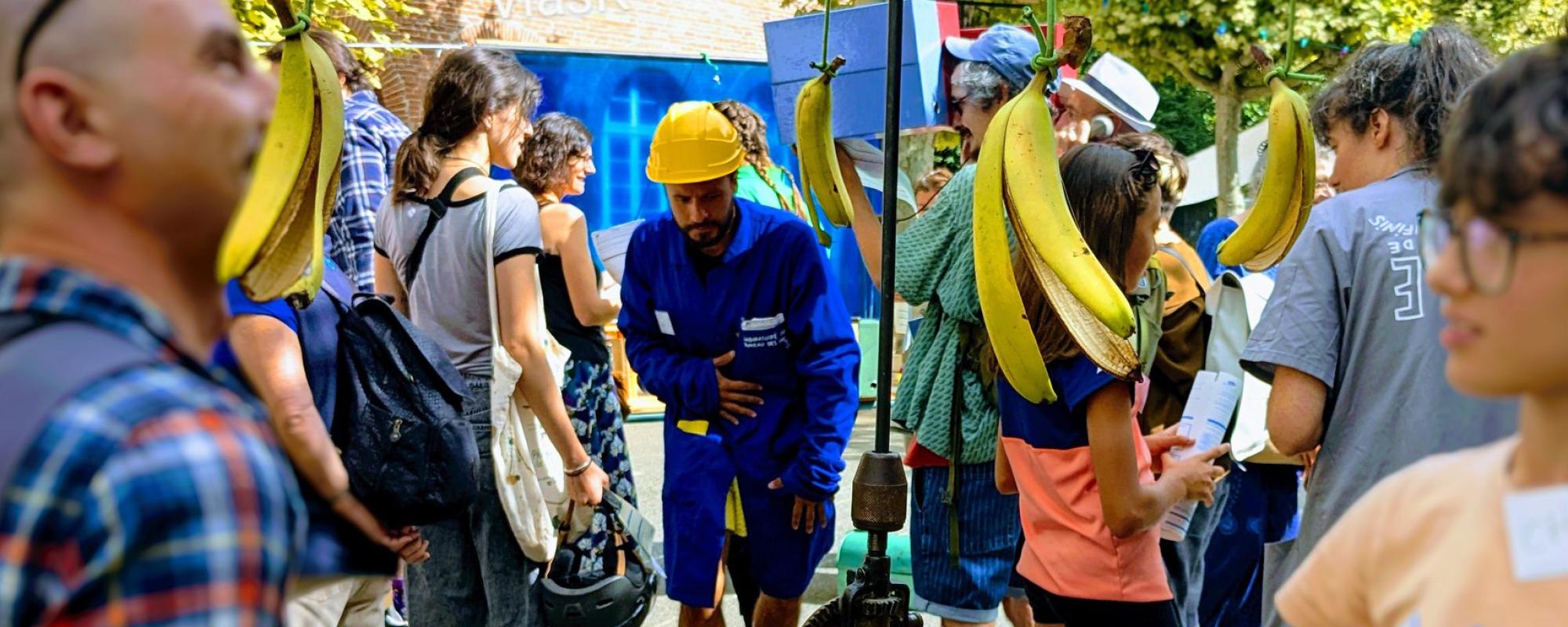 Des personnes dehors, un garçon tient une cannes à pêche avec des bananes au bout. Au milieu se tient un comédien avec une blouse bleue et un casque de chantier.