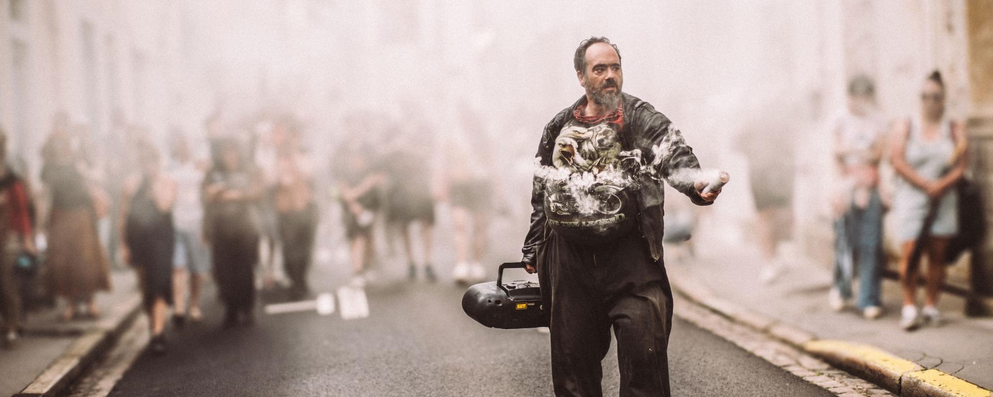 Un homme se tient au milieu de la rue. Il tient dans une main une grosse radio CD et dans l'autre un fumigène blanc. La rue est enfumée et un groupe de personne du public se tient à l'arrière, dans la fumée.