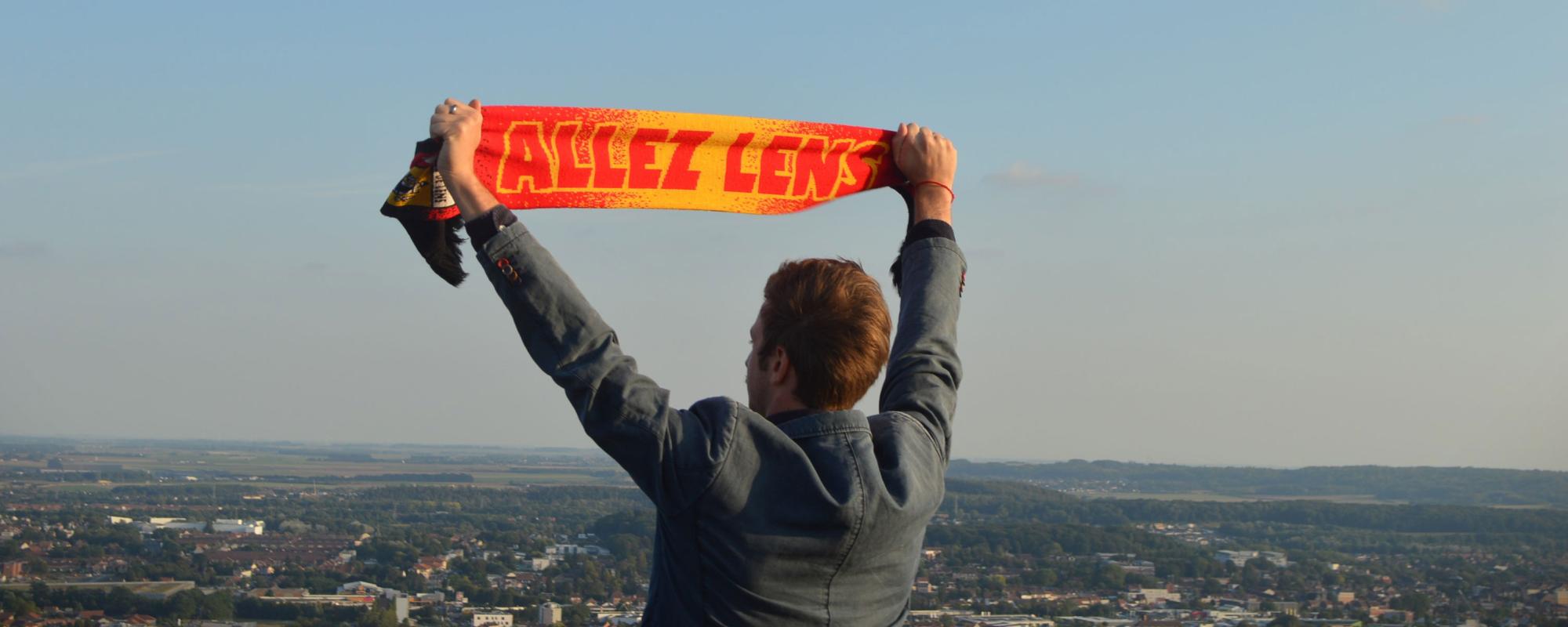 © Raphaëlle Viau Le comédien se tient de dos et brandit une écharpe de foot rouge et jaune "ALLEZ LENS". Il regarde l'horizon et les villes environnantes en contrebas.