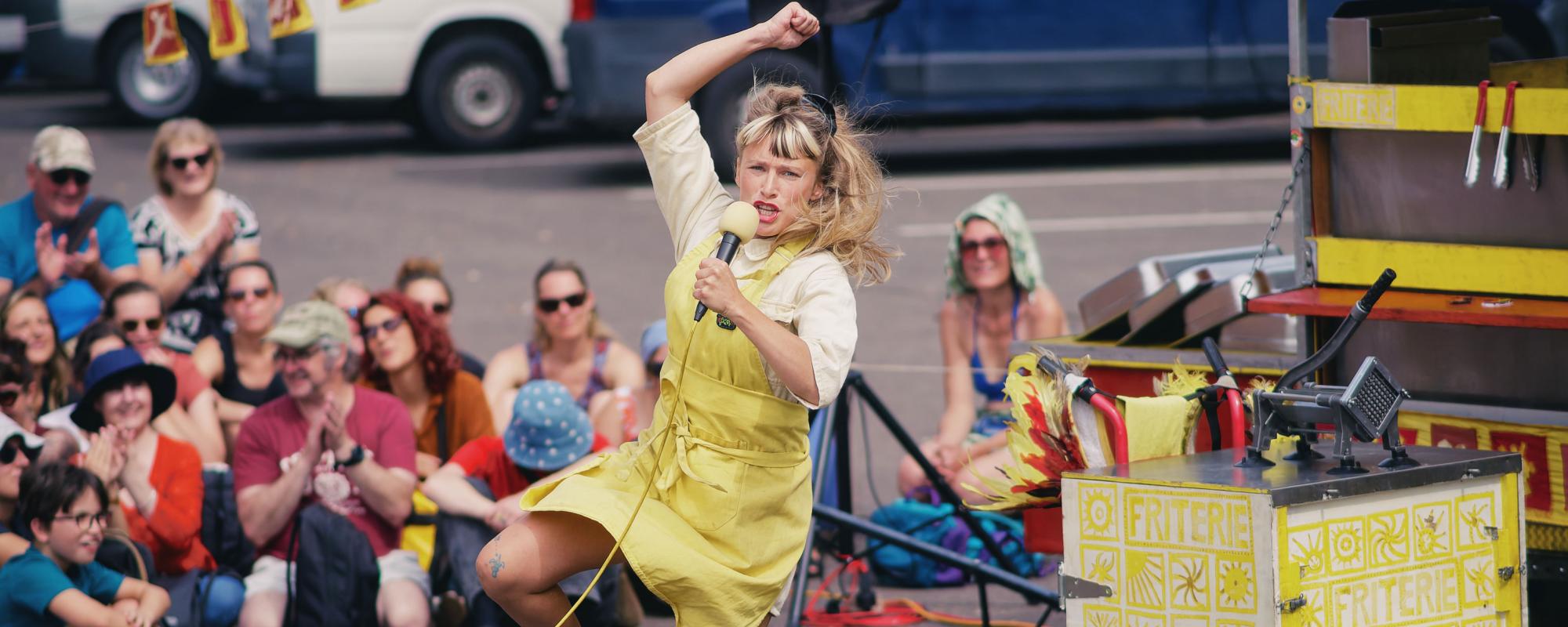 La comédienne porte un tablier jaune et tient un micro dans sa main gauche. Elle semble en train de sauter, un bras en l'air et une jambe pliée. Derrière elle, un coupe-frites ambulant, décoré avec des motifs jaunes et l'inscription "Friterie". À l'arrière, le public est assis par terre, légèrement flouté.