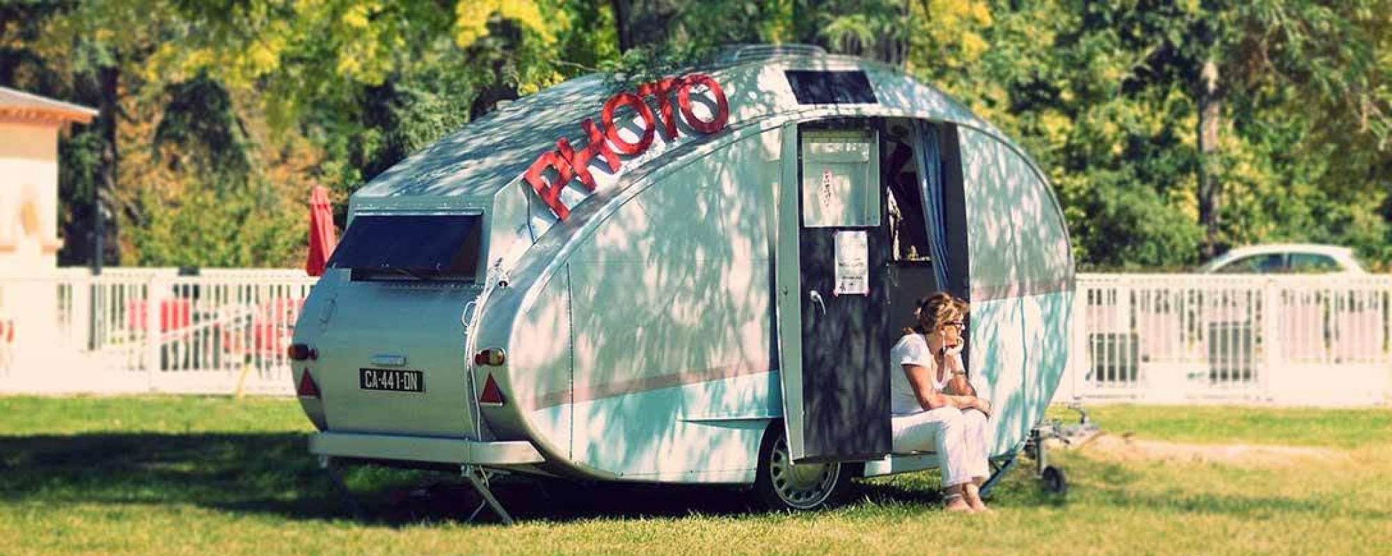 Caravane vintage dans une étendue d'herbe avec un lettrage "PHOTO" posé dessus. Une femme est assise sur le rebord de la porte.
