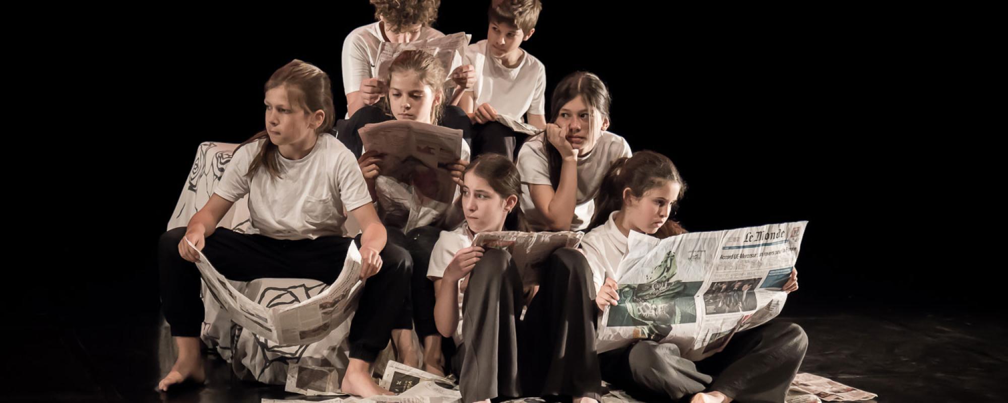 Neufs enfants sont assis et assises sur scène et lisent des journaux. Iels portent un t-shirt blanc et un pantalon noir. Des journaux sont éparpillés sur le sol.
