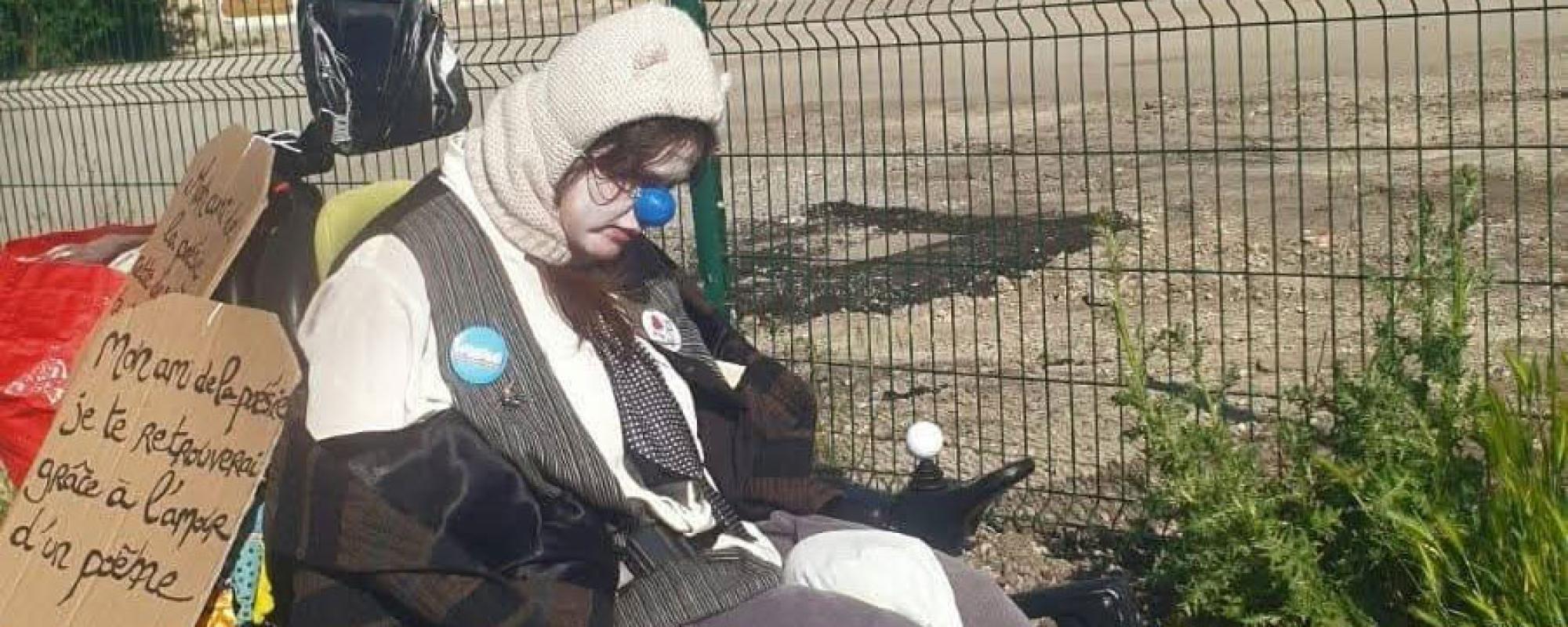Un clown en fauteuil roulant électrique porte une cagoule blanche avec des petites oreilles. Son visage est visible, peint en blanc et il porte un nez bleu en plastique. Au dos de son fauteuil sont accrochées des pancartes en carton.