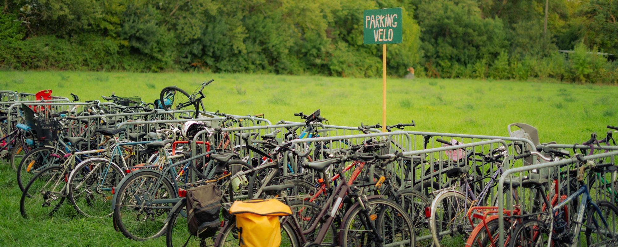 Dans un champ, des vélos sont parqués, attachés à des barrières vauban. Une pancarte en bois colorée indique "PARKING VELO".