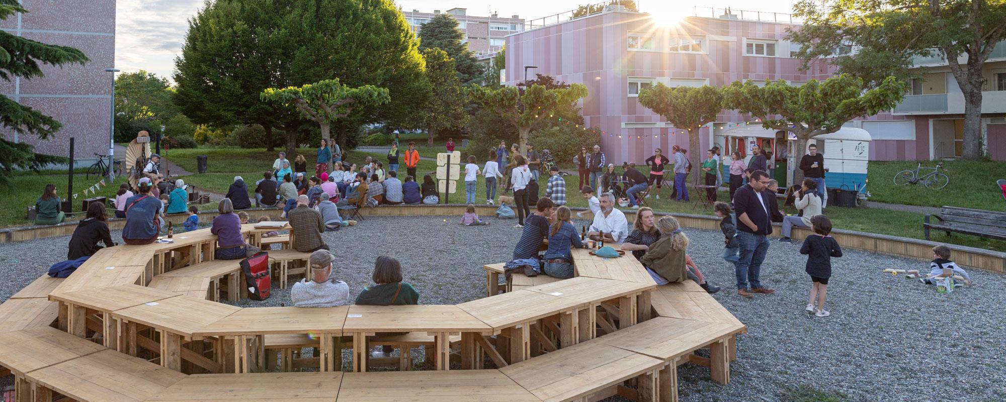 Installation en bois de tables et bancs installés en octogone dans la Cité Rose/Résidence St. Agne de Ramonville. Des personnes sont assises et des enfants jouent autour pendant un concert.