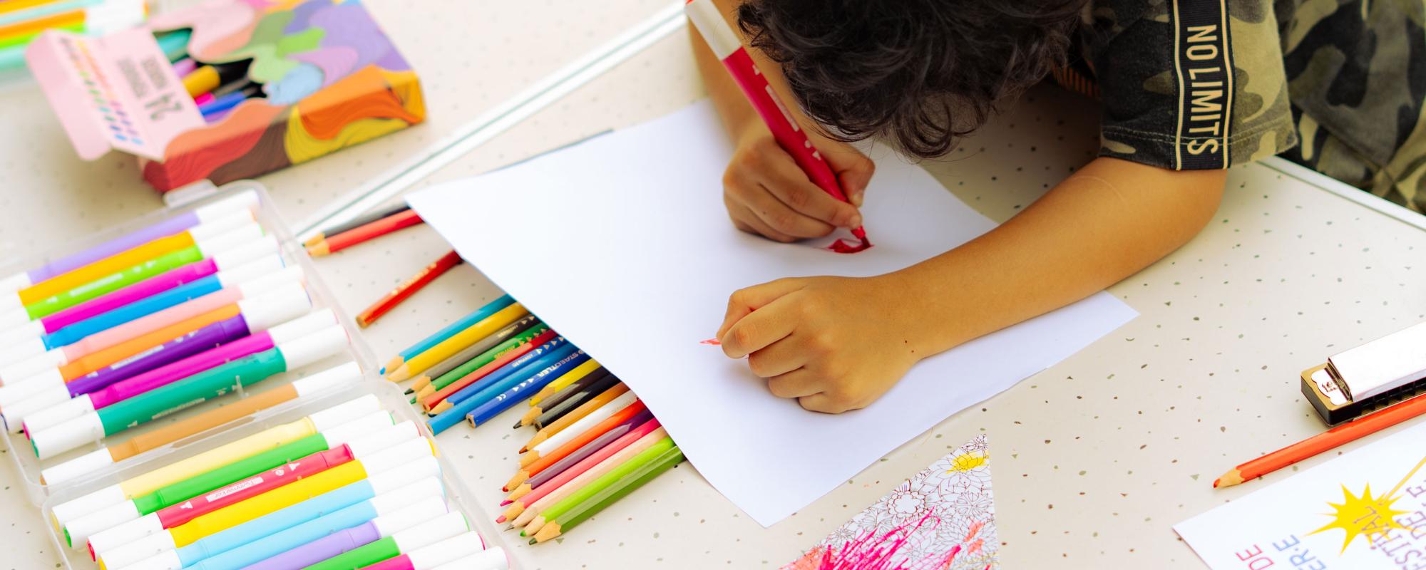 Un enfant dessine à une table. Il y a plein de crayons et de feutres.